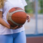 Crop unrecognizable woman in activewear on basketball court with ball near fence in daylight
