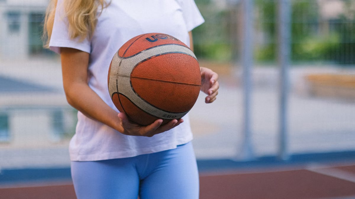 Crop unrecognizable woman in activewear on basketball court with ball near fence in daylight